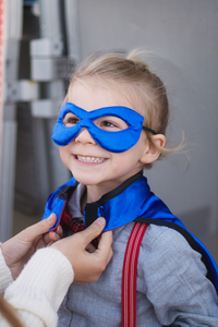 Image of child wearing eye mask as an adult (with only their hands in frame) velcros their cape together.  Unrelated to the costume, the kid is also wearing read suspenders.  