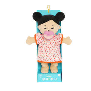 Wee Baby Stella Light Beige with Black Buns in packaging.  She is wearing an orange dress with oranges on it and a purple pacifier is in her mouth.  